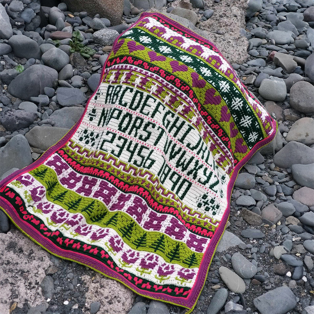 Rosina Plane Lakenham Mosaic Sampler CAL Haakpakket - Bright deken gedrapeerd over stenen op het strand